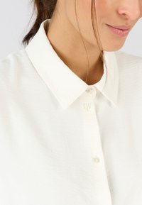 Weißes Hemd mit Knöpfen und einem strukturierten Kragen sowie einer leicht zerknitterten Textur. Es verfügt über kleine, klare Knöpfe und ein minimalistisches Design.
