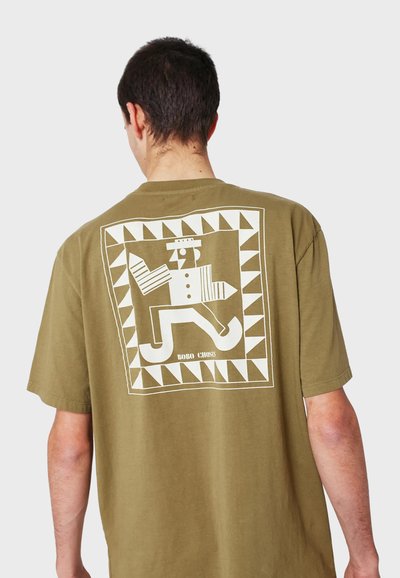 Olivově zelené bavlněné tričko s velkým bílým grafikou na zadní straně, která má geometrický rámeček a design postavy. Krátké rukávy, uvolněný střih.