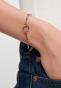 Guldtonet armbånd med sammenkoblede papirclips-formede og cirkulære elementer, vist på et håndled mod en denim-baggrund.