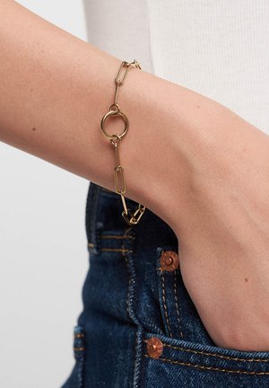 Goldfarbener Armreif mit verbundenen, papierclipförmigen und kreisförmigen Elementen, am Handgelenk vor einem Denim-Hintergrund präsentiert.