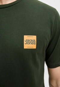 Dunkelgrünes T-Shirt aus Baumwolle mit einem quadratischen orangefarbenen Logo und dem Text "Jack & Jones" in Schwarz, platziert auf der linken Brustseite. Kurze Ärmel.