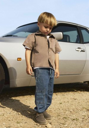 Barn iført en lysebrun kortærmet skjorte med knapper, blå denim cargobukser og brune slip-on sko står nær en sølv bil.