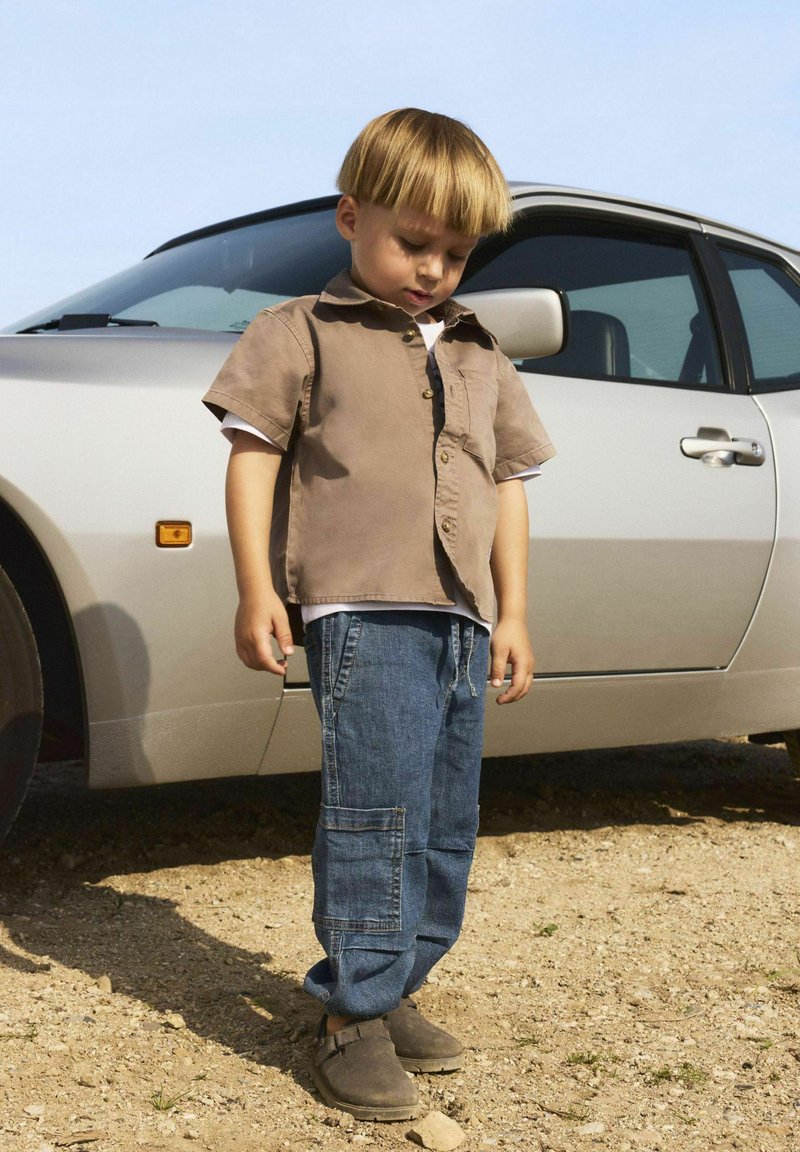Un enfant portant une chemise à manches courtes marron clair, un pantalon cargo en denim bleu et des chaussures slip-on marron se tient près d'une voiture argentée.