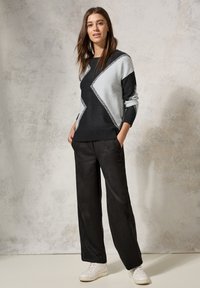 Pull en tricot noir et gris avec un motif géométrique, associé à un pantalon large noir et des baskets blanches sur un fond clair.