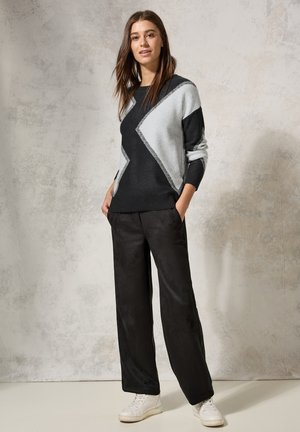 Pull en tricot noir et gris avec un motif géométrique, associé à un pantalon large noir et des baskets blanches sur un fond clair.