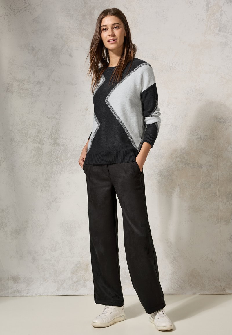 Schwarzer und grauer Strickpullover mit geometrischem Design, kombiniert mit schwarzen weit geschnittenen Hosen und weißen Sneakers vor einem hellen Hintergrund.