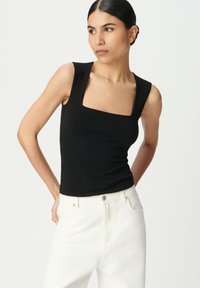 Mujer con pelo oscuro en un moño bajo, vistiendo una blusa sin mangas de cuello cuadrado negra y jeans blancos de cintura alta, posando con las manos detrás de la espalda.