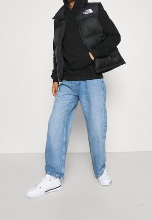 Sort puffervest med hætte over en sort hættetrøje, kombineret med løstsiddende lyseblå jeans og hvide sneakers. Enkel design, ingen synlige mønstre.