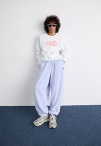 Sweatshirt branca com o logótipo "Nike" em rosa, combinado com umas calças de treino largas em roxo claro e ténis cinzentos com padrão. Óculos de sol escuros usados.