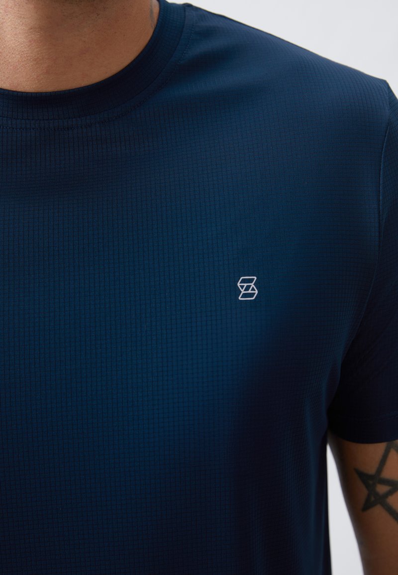 Camiseta de color azul marino que absorbe la humedad, con un patrón texturizado, escote redondo y un pequeño logo blanco en el pecho. Tela suave y mangas cortas.