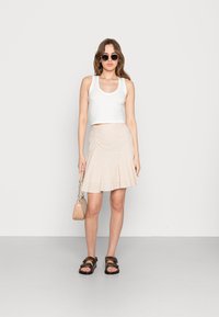 Top cropped branco, saia plissada com padrão xadrez bege e branco, sandálias pretas e uma pequena mala de palha. Os acessórios incluem óculos de sol redondos.