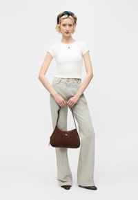 Kvinne iført en hvit kort topp, lysegrå bukser med vide ben, rutete hårbånd, brune sko og holder en brun skulderveske foran en hvit bakgrunn.