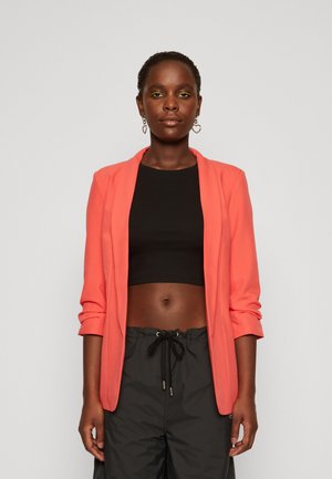 Blazer corallo con maniche lunghe, apertura frontale e texture liscia, abbinato a un top corto nero e a pantaloni corti neri con coulisse.