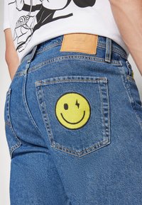Blue denim jeans featuring a large yellow embroidered smiley face on the back pocket, with a leather patch on the waistband.