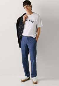 Homme portant un t-shirt blanc "PEPE JEANS", un pantalon bleu, des baskets blanches, tenant une veste en denim foncé sur une épaule, debout devant un fond uni.