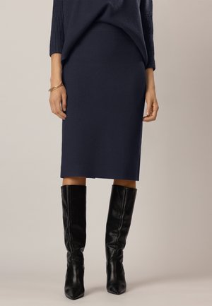 Femme portant une jupe bleu marine longueur genoux, des bottes en cuir noires montantes aux genoux et un haut à manches longues foncé avec un bracelet au poignet gauche.