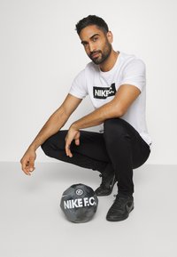 Weißes Nike T-Shirt mit schwarzem Logo, schwarze Trainingshose und schwarze Fußballschuhe. Ein schwarz-weißer Nike Fußball liegt auf dem Boden.