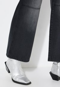 Stivali ankle metallici argento con punta a punta e tacco a blocco, abbinati a pantaloni a gamba larga in denim scuro con dettagli di cucitura visibili.