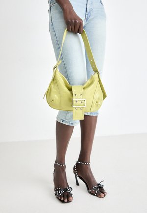 Žlutá perforovaná kabelka s detaily přezky a nastavitelným popruhem, spárovaná s denimovými šortkami a černými sandály na vysokém podpatku s mašlemi.