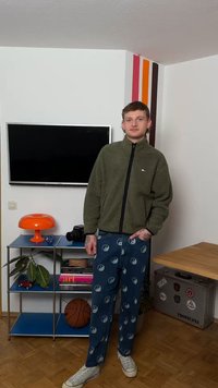 Jeune homme debout dans une pièce avec un coin de mur rayé, portant une veste verte zippée, un pantalon à motif yin-yang, des baskets blanches, près d'un ballon de basket et d'une lampe orange.