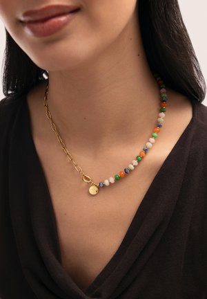 Primer plano de una persona que lleva un collar de cuentas multicolor con una cadena de oro y un colgante redondo de oro, vestida con una prenda oscura.