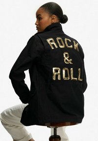 Chaqueta negra con cuello, que presenta letras doradas de lentejuelas que dicen "ROCK & ROLL" en la parte posterior. Tejido suave con un corte relajado.