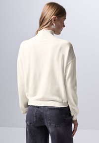 Pullover bianco in maglia con collo alto e vestibilità comoda, caratterizzato da polsini e orlo a costine, abbinato a jeans grigio scuro.