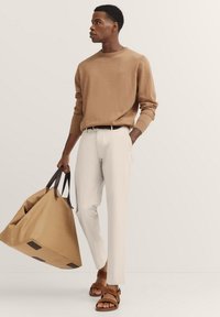 Beiger Strickpullover, weiße Hose, braune Ledersandalen und eine große, sandfarbene Tragetasche mit dunklen Akzenten. Einfaches, modernes Design, das auf neutralen Farben basiert.