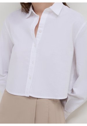 Weißes, langärmeliges Cropped-Shirt mit Kragen und Knöpfen, getragen über beigen, hochgeschnittenen, plissierten Hosen an einer Person.