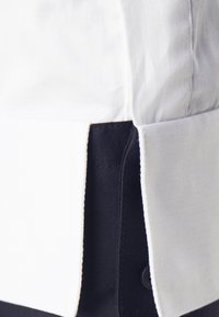 Weißes und marineblaues Hemd mit Kragen und kontrastierender Seitennaht. Glatte Textur, sichtbare Nähte und ein Knopfdetail entlang des marineblauen Abschnitts.