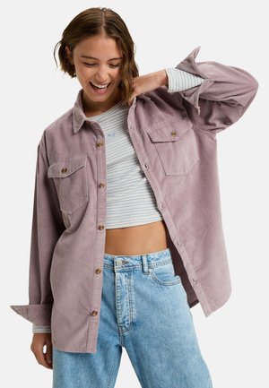 Lichtpaarse ribfluwelen shirt met twee borstzakken, knopen aan de voorkant en opgerolde mouwen, gelaagd over een gestreept cropped top en blauwe spijkerbroek.
