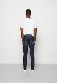 Man som bär en vit t-shirt, mörkblå jeans och vita sneakers, stående vänd mot en enkel ljusgrå vägg.