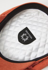 Căciulă din material portocaliu, cu un interior moale, căptușit de culoare albă. Prezentă un eticheta neagră cu textul "Brixton" și un design de logo. Decupată în negru.