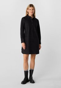 Schwarzes langärmliges Kleid mit Button-Down-Kragen, geradem Schnitt, strukturiertem Stoff und Seitentaschen, kombiniert mit schwarzen kniehohen Stiefeln.