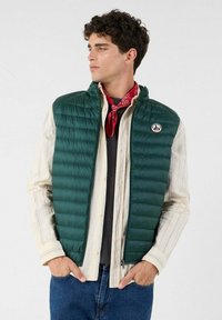 Gilet imbottito verde con texture trapuntata, chiusura a zip e logo. Indossato sopra una camicia a righe, abbinato a jeans blu e una bandana rossa.