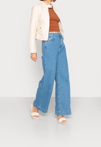 Světle hnědá kožená bunda přes hnědý top, doplněná volnými světle modrými denimovými širokými kalhotami a bílými sandály na blockovém podpatku se zlatým páskem.