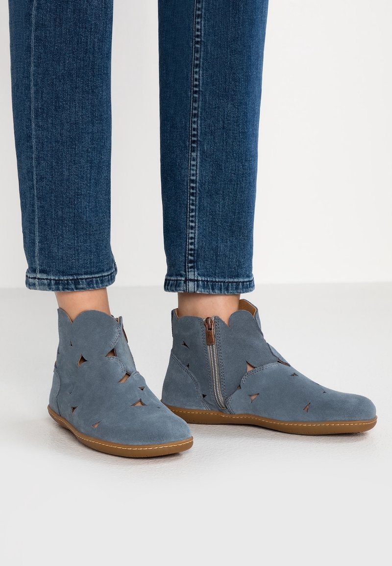 Bottines en daim bleu avec des motifs découpés et des fermetures éclair latérales, portées avec un jean en denim bleu.