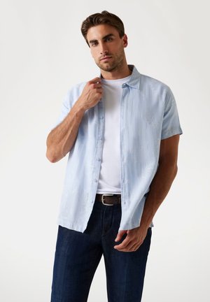 Camisa de manga corta azul claro con un sutil patrón floral, usada sobre una camiseta blanca, combinada con pantalones de mezclilla oscuros y un cinturón marrón.