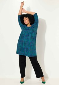 Tunika mit grünem und lila Muster, drei Viertelärmel, kombiniert mit schwarzen Hosen und grünen Schuhen, im lockeren, fließenden Design.