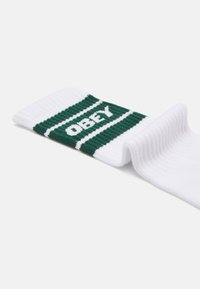 Λευκές κάλτσες μέχρι τον αστράγαλο με πράσινες οριζόντιες ρίγες που φέρουν τη λέξη "OBEY" σε λευκό, υφασμένο ύφασμα, και σχέδιο ribbed στο γιακά.