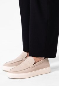 Beige Wildleder Slipper mit einer runden Zehenpartie, dekorativer Naht und einer weißen Gummisohle, kombiniert mit schwarzen, cropped Hosen.