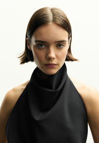 Schwarzes ärmelloses Oberteil mit drapierter Kragenform, glatter Textur und minimalistischer Gestaltung. Das Modell hat kurzes Haar und neutrale Make-up auf einem weißen Hintergrund.