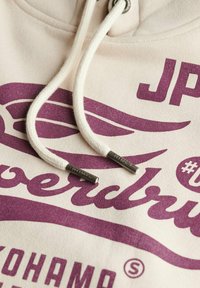 Crème hoodie met paarse grafische tekst, met witte trekkoorden en zilveren hardwareaccenten. Zachte textuur en casual ontwerpdetails.