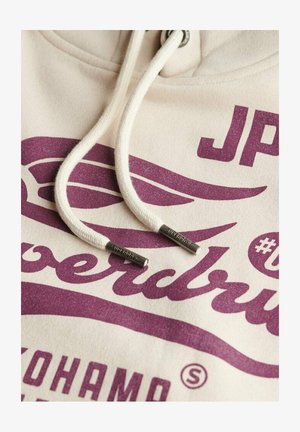 Creme hoodie med lilla grafisk tekst, med hvide snore og sølv hardware-detaljer. Blød tekstur og afslappet design.