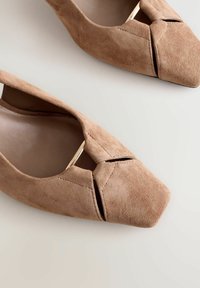 Zapatos de tacón de gamuza color marrón claro con punta afilada y diseño recortado, que presentan un acento metálico. Textura suave con un acabado mate sutil.