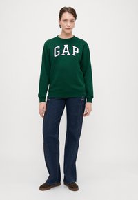 Grønn hettegenser med hvit "GAP" skrift, kombinert med mørkeblå utvidede jeans. Jeansene har side-d stitching og er brukt med brune sko.
