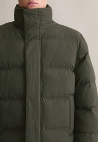 Geacă puf verde cu un guler înalt, textură matlasată, închidere frontală și două buzunare laterale. Material moale și ușor.
