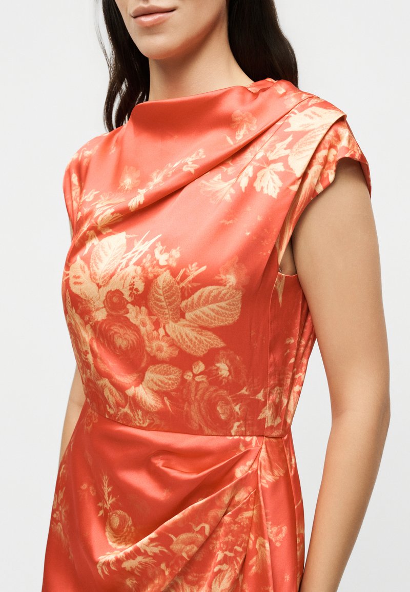 Woman wearing sleeveless coral satin dress with golden floral print and draped neckline, shown from shoulders to waist.