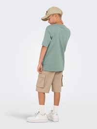 Lys grøn kortærmet t-shirt, beige cargo shorts med lommer, hvide sneakers og en beige kasket båret baglæns.