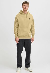 Beige hettegenser med kenguru-lomme, snøring i hetten, og liten logo-detalj på brystet, kombinert med mørke jeans og hvite joggesko.